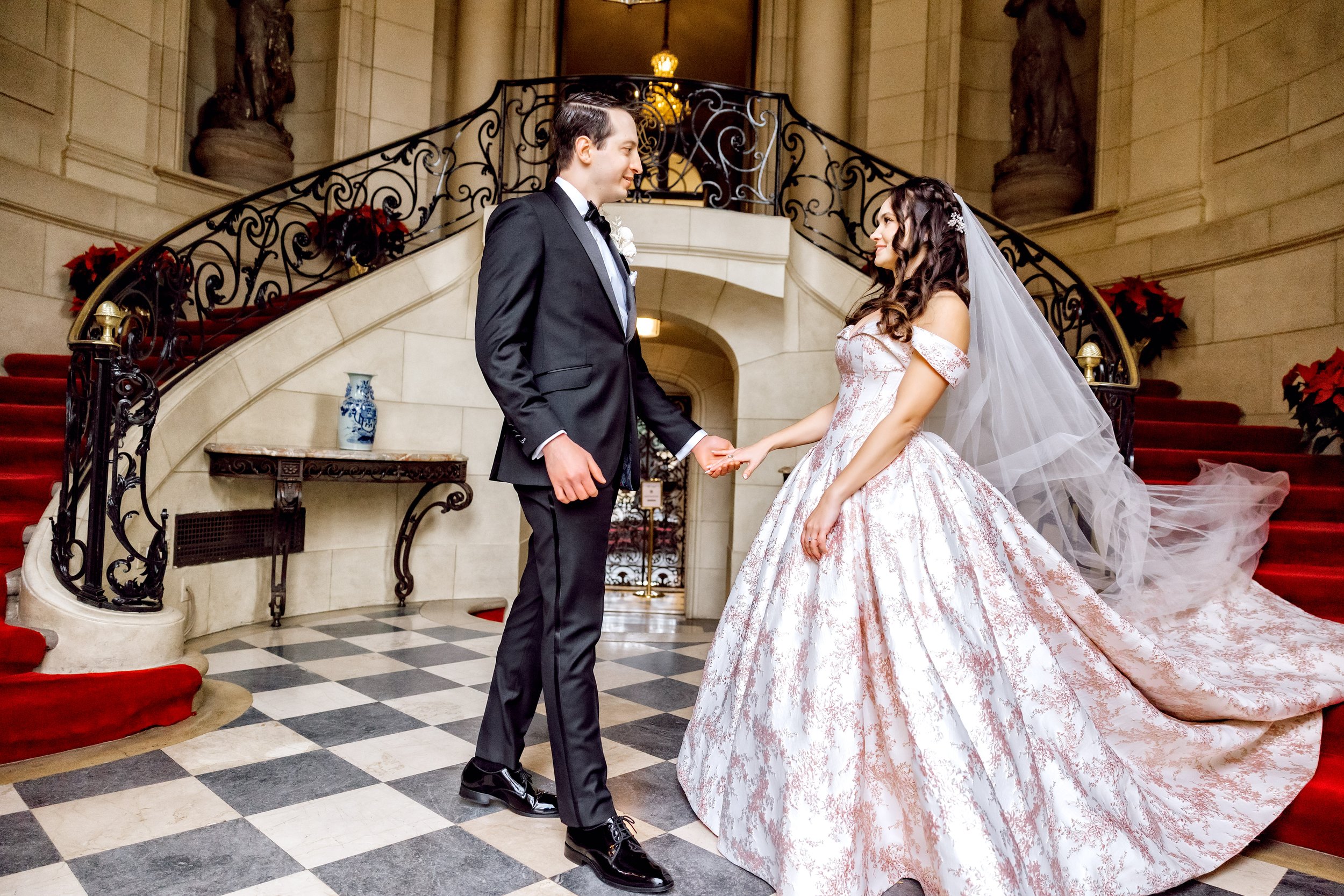 A bride and groom holding hands on a staircase at their wedding, with the bride in a white gown and veil and the groom in a black tuxedo, in an elegant hall with red carpet and decorative elements.