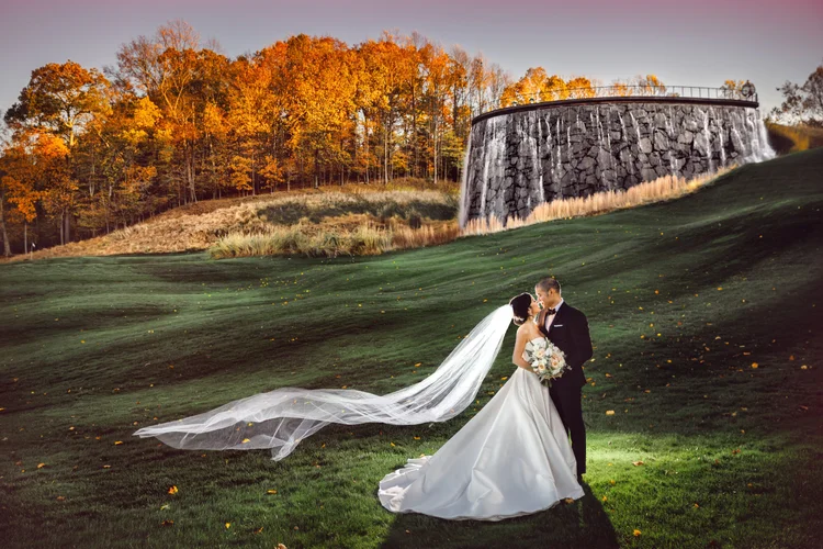 A bride and groom standing on a grassy field during sunset, with the bride wearing a white wedding gown and veil and holding a bouquet of flowers, while the groom wears a black tuxedo. The bride's veil is flowing behind her. In the background, there 