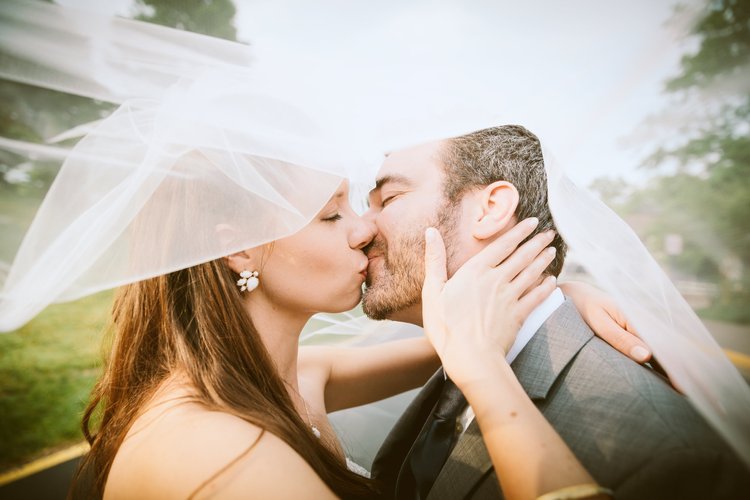 A bride and groom are kissing beneath a white veil outdoors during daytime.