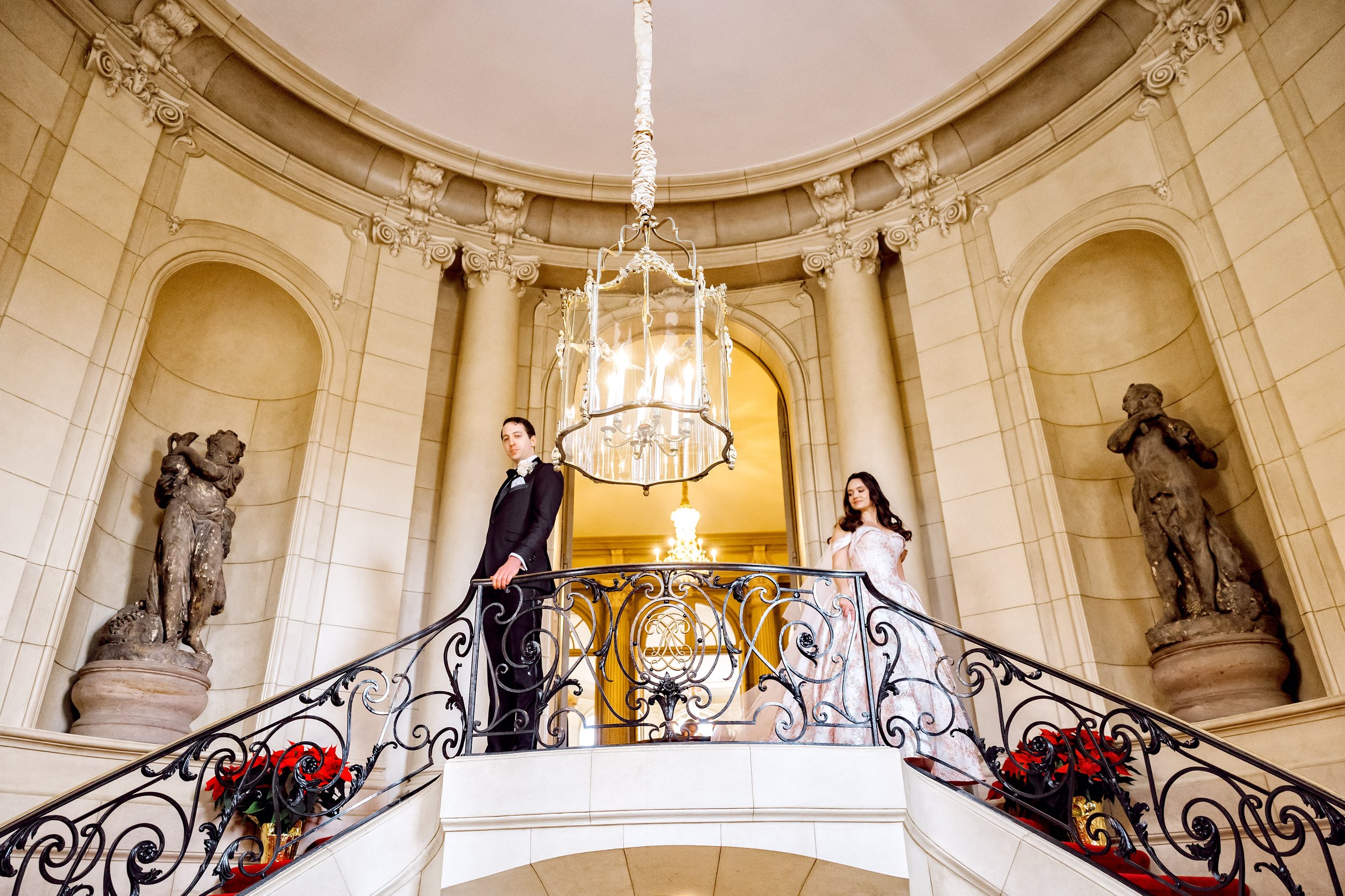 A man and woman in formal attire standing on a grand staircase with ornate black wrought iron railing inside a luxurious building. The man is dressed in a tuxedo, and the woman is in a white wedding gown. The background features classical sculptures,