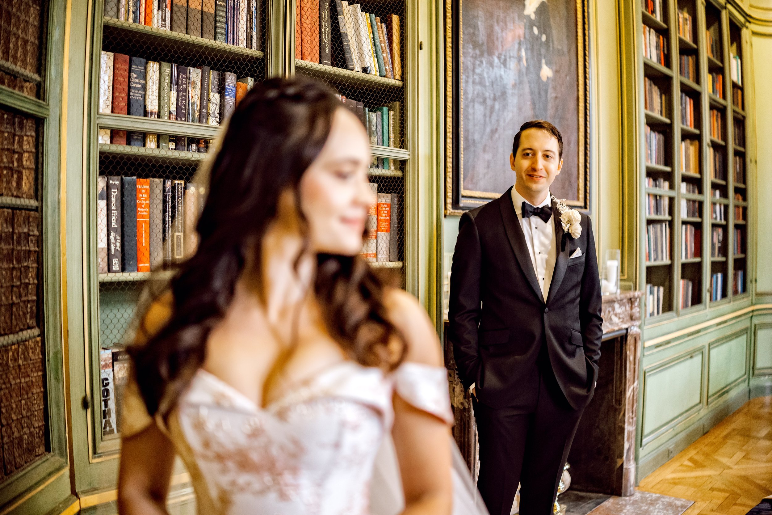 A bride and groom at their wedding, standing in a vintage library with bookshelves and a fireplace in the background. The groom is smiling while looking at the bride, who is slightly out of focus.