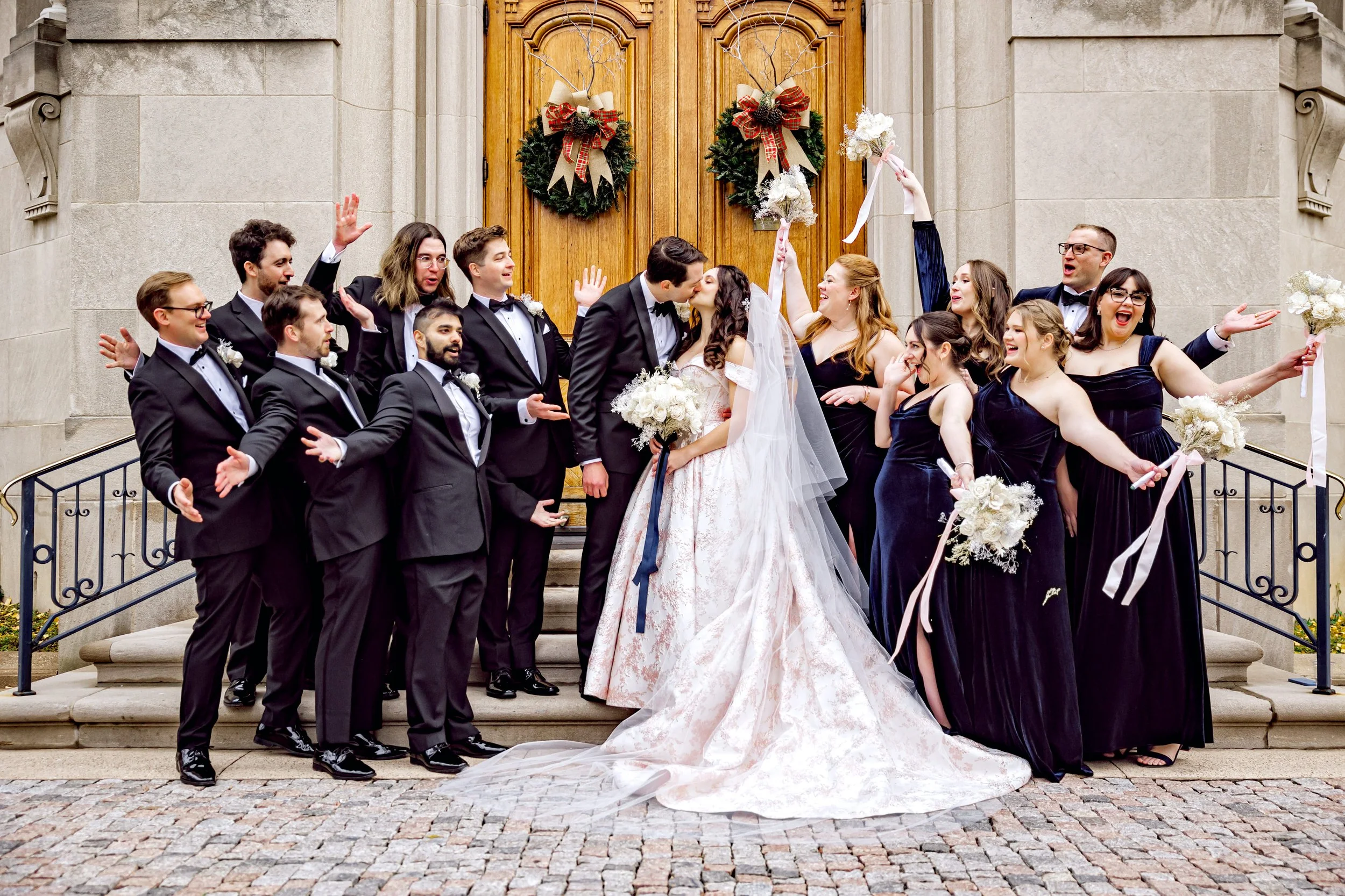 A wedding celebration with a bride and groom kissing in front of a church with wedding party members around them, some holding bouquets and others raising their arms, on steps decorated with wreaths and Christmas bows.