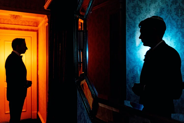 Silhouettes of two men in tuxedos facing each other in a dimly lit room with colorful lighting, one on the left with orange hues and one on the right with blue hues, separated by a wall with framed pictures.