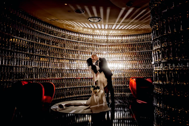 A bride and groom share a kiss in a luxurious, dimly lit room with walls covered in champagne bottles. The bride holds a bouquet, and the groom wears a tuxedo. The room has modern decor with red accents and reflective flooring, creating a romantic at