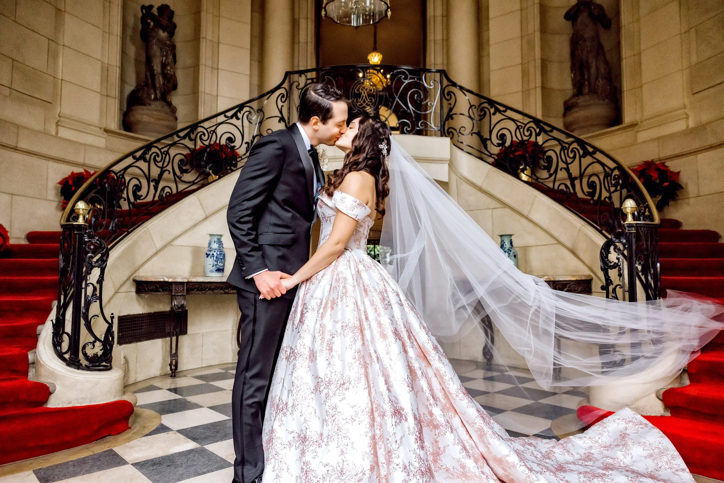 A bride and groom sharing a kiss in a grand staircase hall, with the bride wearing a white wedding gown and veil, and the groom wearing a black tuxedo.