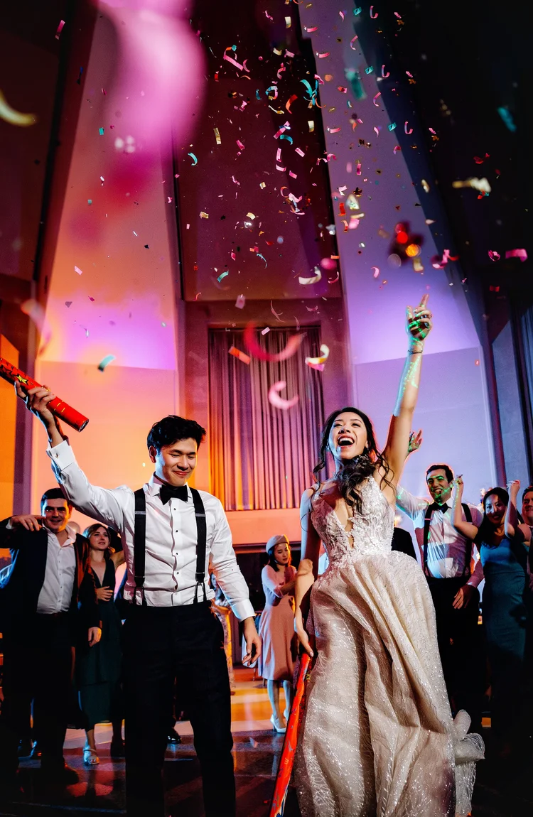 A bride in a wedding gown celebrating at her wedding reception surrounded by guests with confetti falling from above.