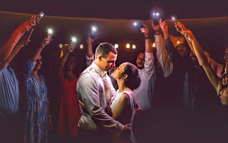 A bride and groom dancing closely at their wedding reception, surrounded by friends who are taking photos with their phones illuminated with flash, under warm ambient lighting.