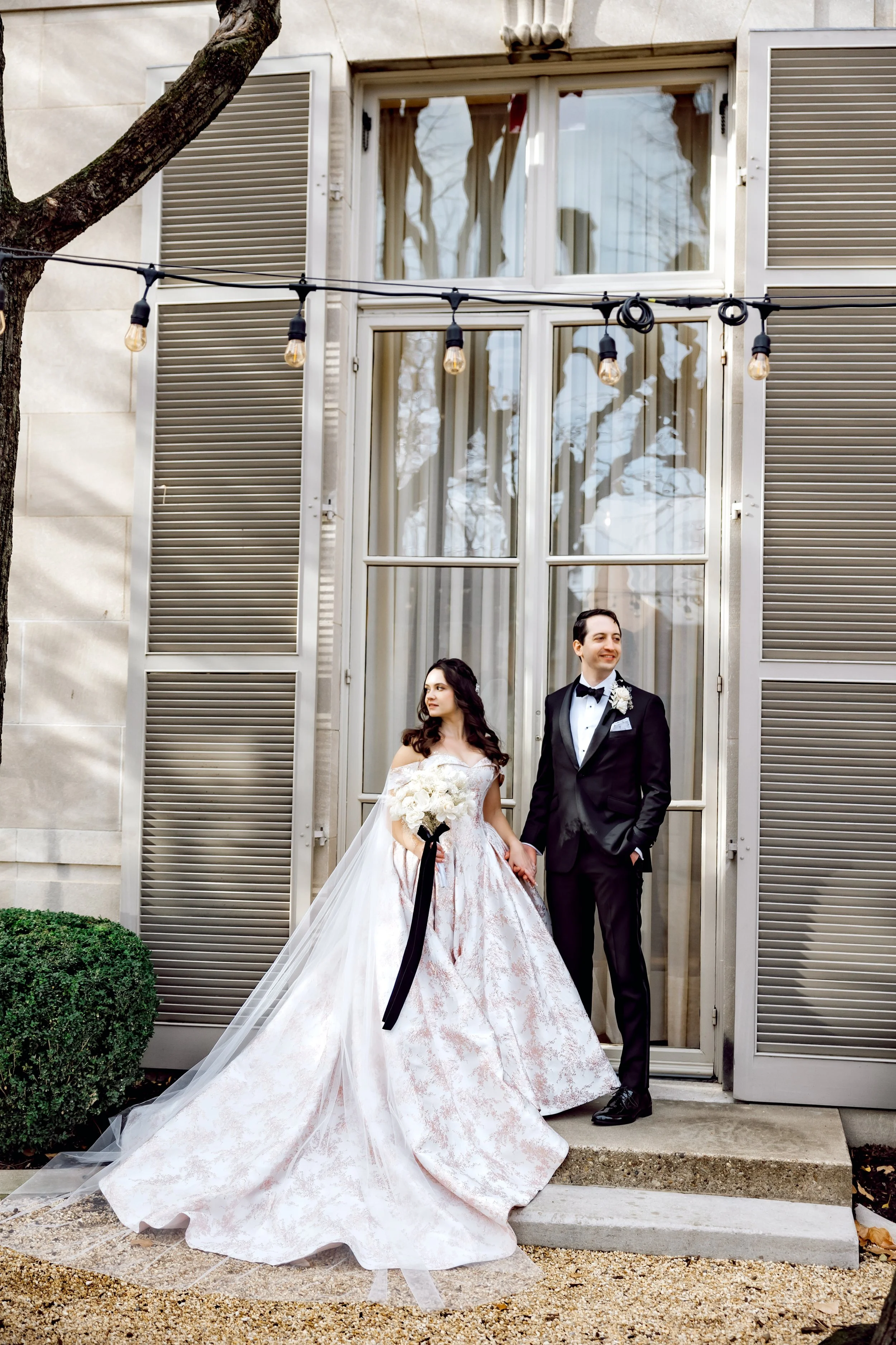 A bride and groom stand outside a building, holding hands. The bride wears a white wedding gown with a full skirt and petal pattern, holding a bouquet of white flowers with a black ribbon. The groom is dressed in a black tuxedo with a bow tie and bou