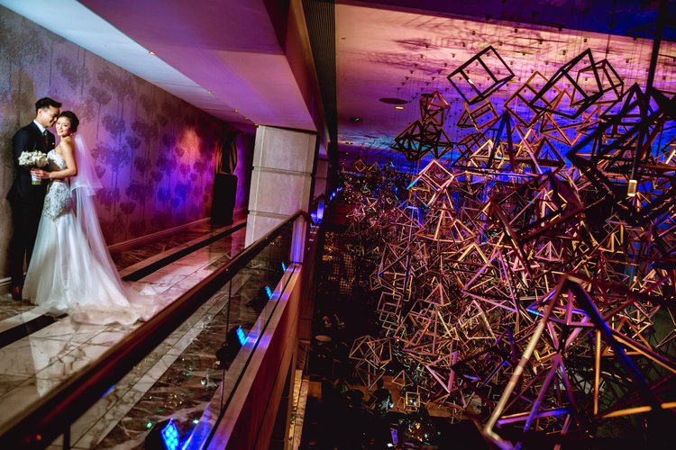 A bride and groom in wedding attire standing on a balcony during their wedding celebration. The background features elaborate colorful geometric light installations and dim, decorative lighting.
