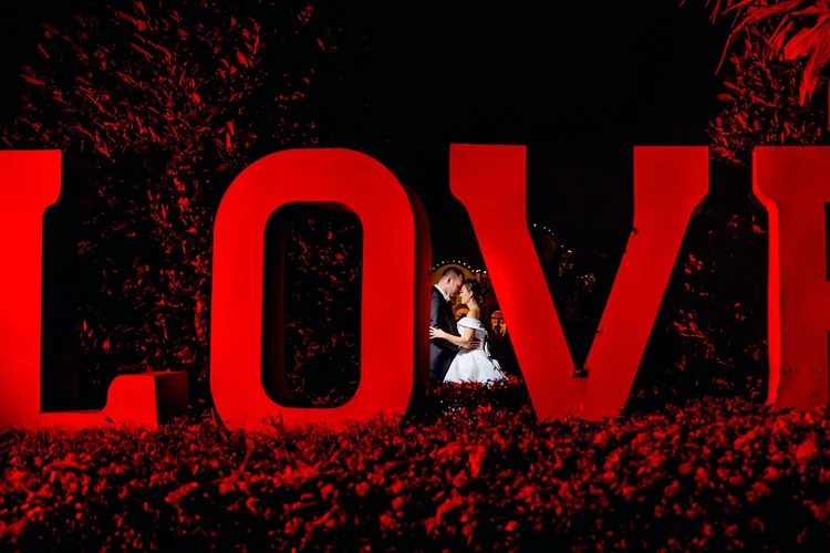 A couple in wedding attire kissing inside a large red 'LOVE' sign at night, with the illumination highlighting them against a dark background.