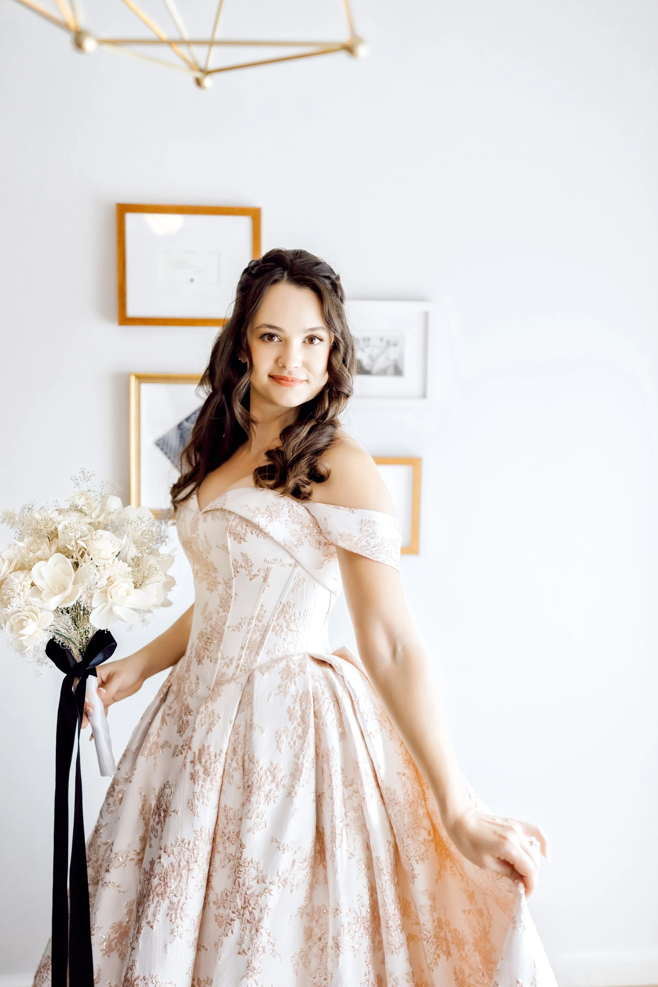 A woman in an off-the-shoulder, vintage-style, peach-colored wedding dress with floral patterns, holding a bouquet of white flowers with black ribbon, standing indoors against a white wall with framed pictures.