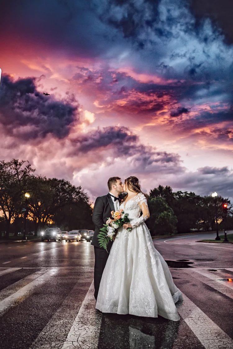 A newlywed couple dressed in wedding attire stands on a crosswalk during sunset. The bride is holding a bouquet of flowers, and they are leaning in close, about to kiss. The sky behind them is colorful with pink, purple, and blue clouds, and cars are