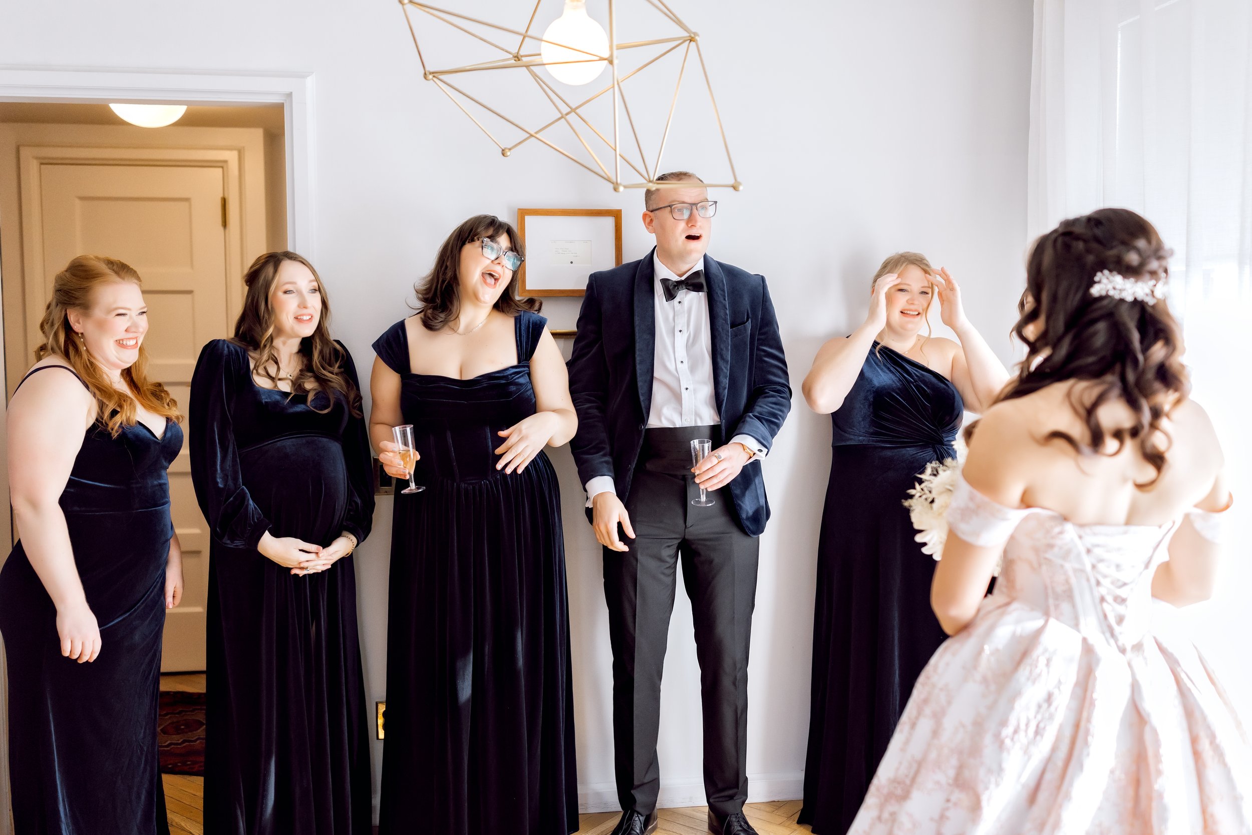 A bride in a white gown with floral details on her shoulders is facing a group of five women and a man, all dressed in formal attire, in a bright room with a geometric gold ceiling fixture.