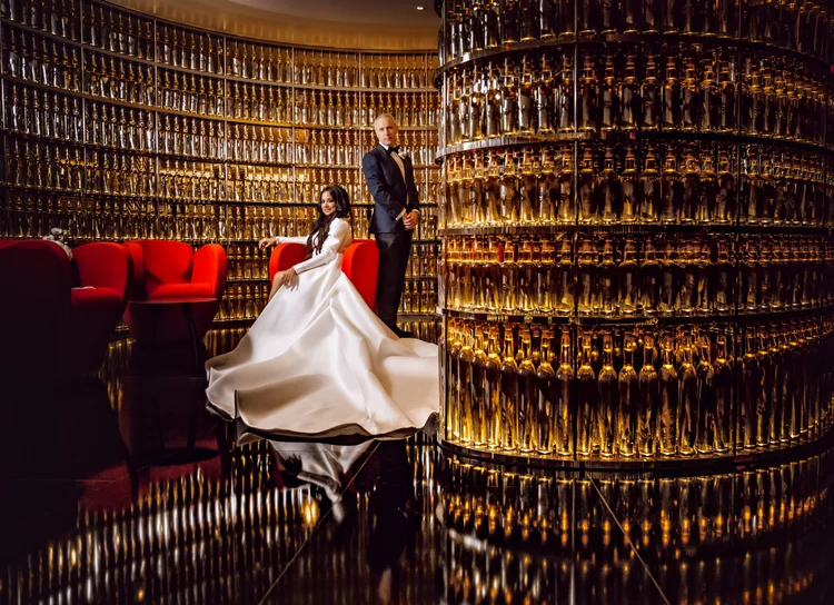 A woman in a white wedding gown seated on a red armchair, and a man in a black tuxedo standing beside her, in a luxurious setting with a circular wall of wine bottles and a reflective black floor.