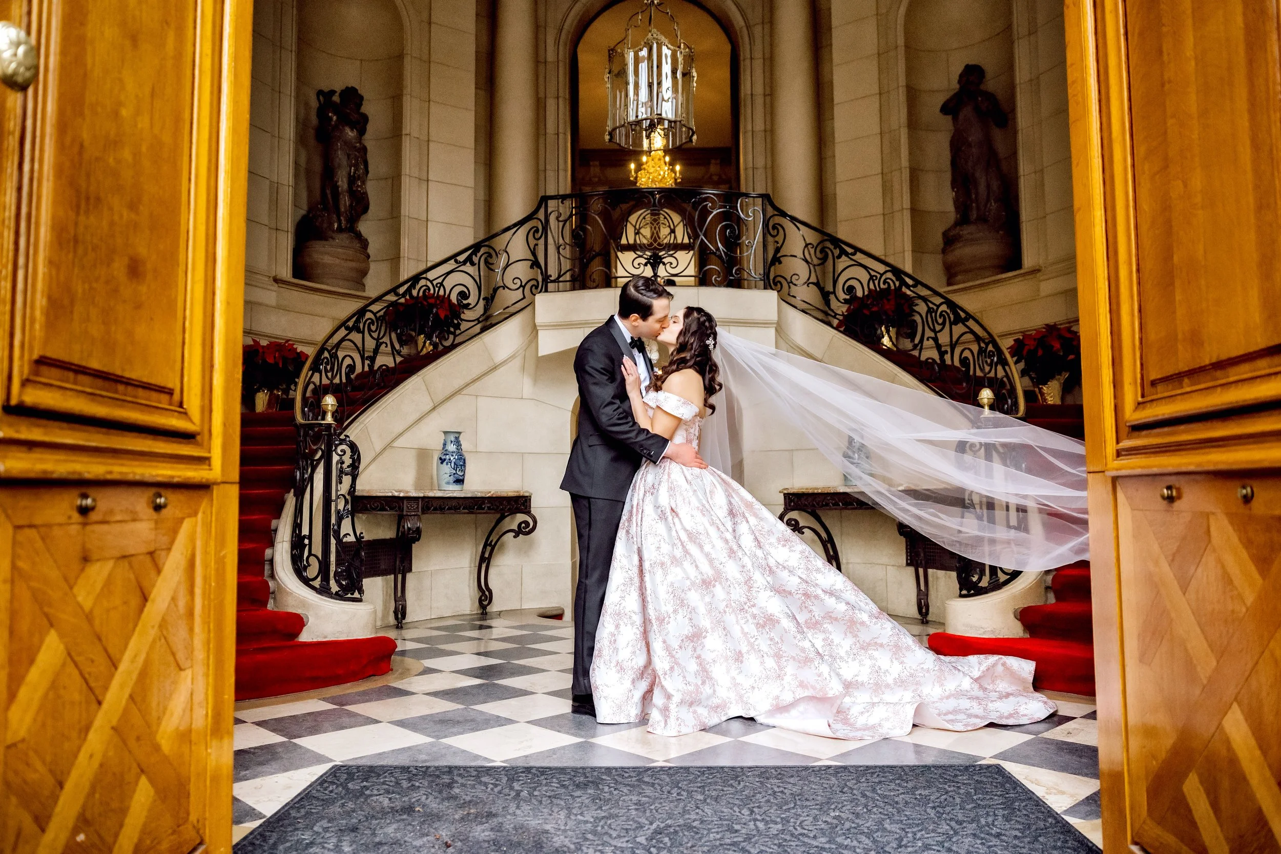A bride and groom kissing in an elegant indoor setting. The bride is in a long, off-the-shoulder white wedding gown with a long veil, and the groom is in a black tuxedo. They stand on a checkered black and white floor in front of a grand staircase wi