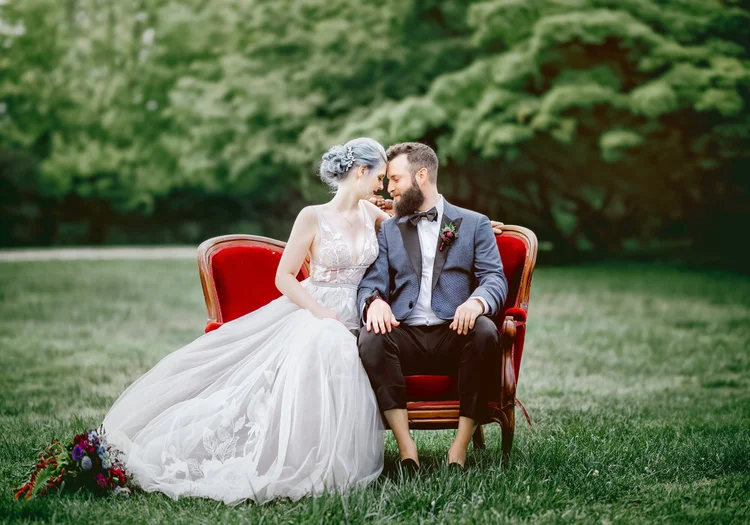 A bride and groom sitting closely on a red velvet vintage sofa outdoors, touching foreheads, with greenery in the background.
