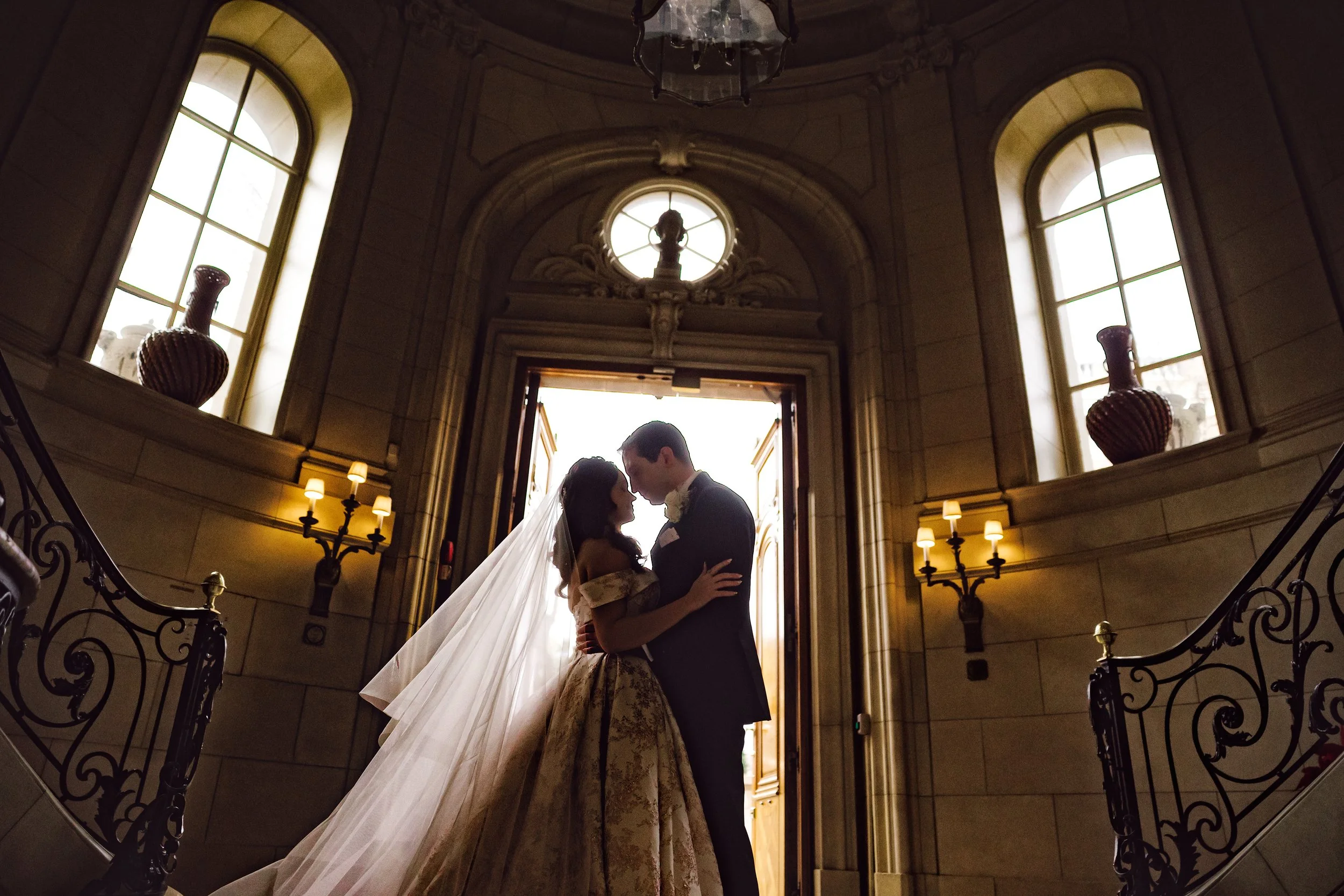 A bride and groom sharing an intimate moment inside a grand, elegant building with high ceilings, large windows, and ornate architecture, backlit by natural light.