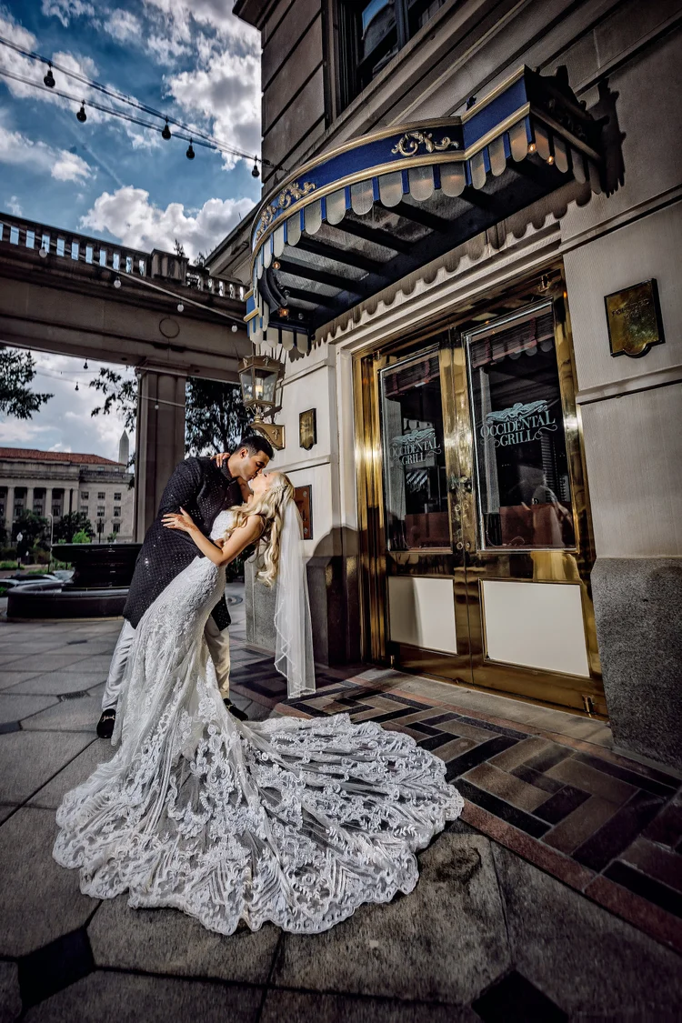 A newlywed couple sharing a kiss outside a restaurant with a gold door, canopy, and string lights overhead, in a city setting.