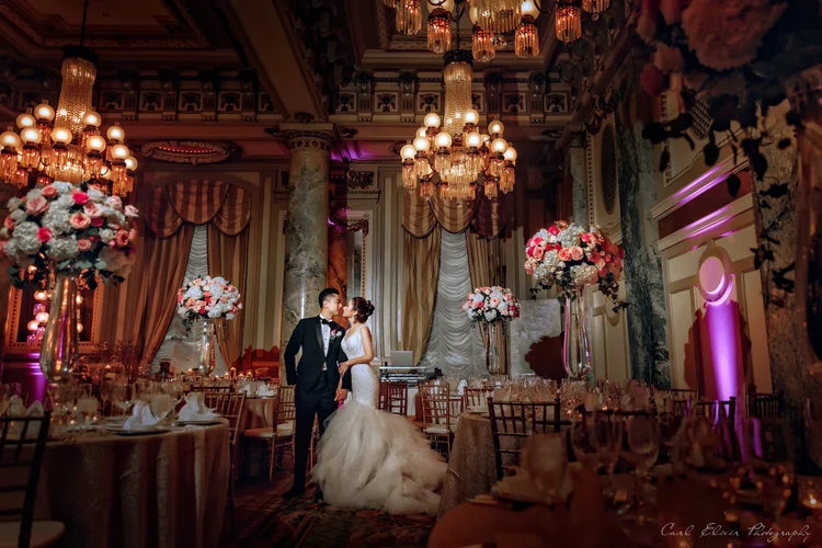 A bride and groom standing close together in an ornate, elegantly decorated ballroom with high ceilings, chandeliers, tall floral arrangements, and draped curtains, sharing a tender moment.