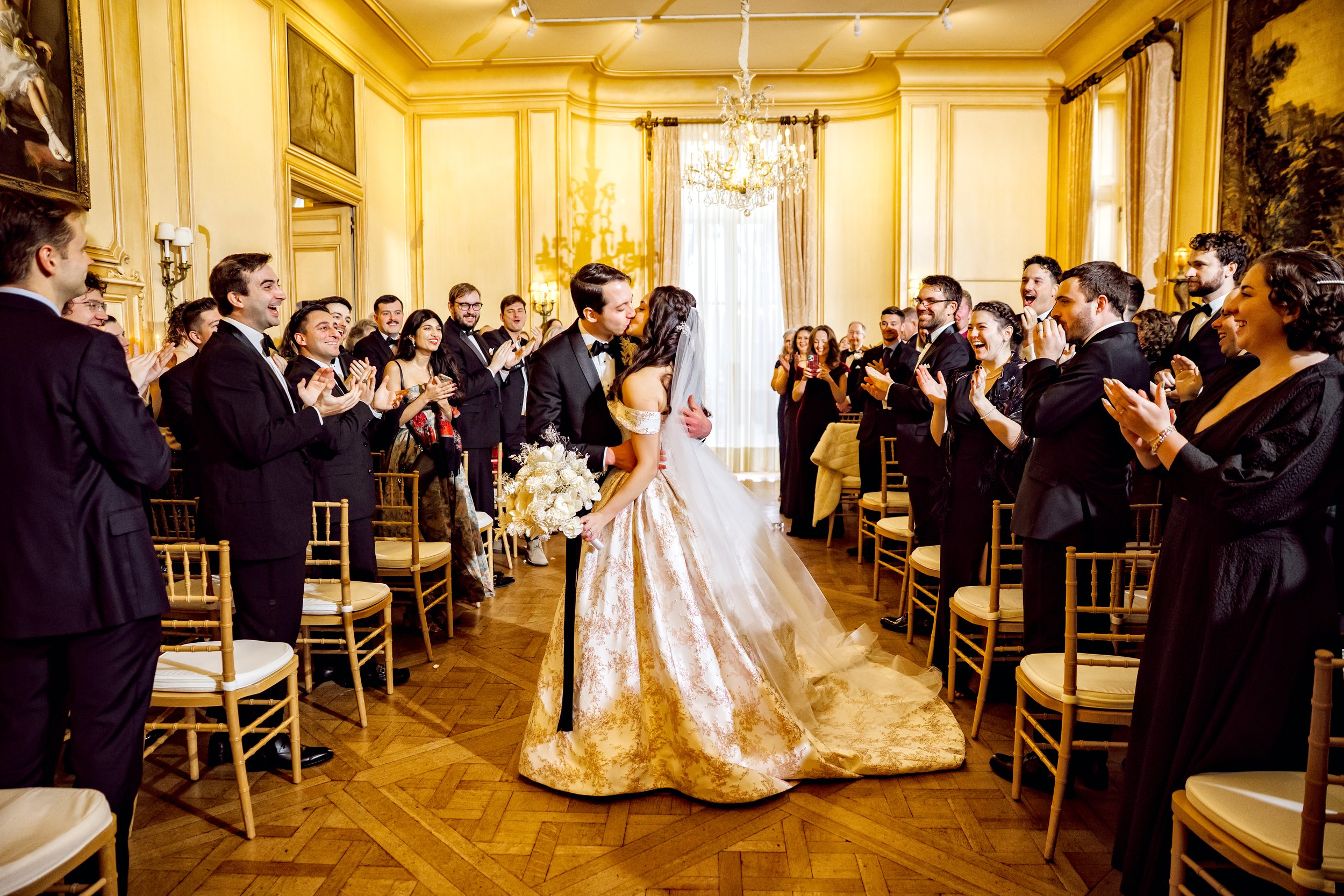 A bride and groom kissing in the center of a room full of wedding guests, with the guests clapping and smiling, in an elegant, well-lit, formal wedding setting with gold-trimmed yellow walls.