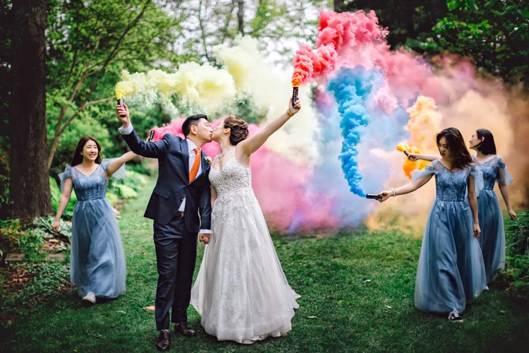 A bride and groom kissing in a wooded outdoor setting while holding colorful smoke grenades, surrounded by bridesmaids holding smoke grenades and releasing colorful smoke in pink, blue, yellow, and orange.