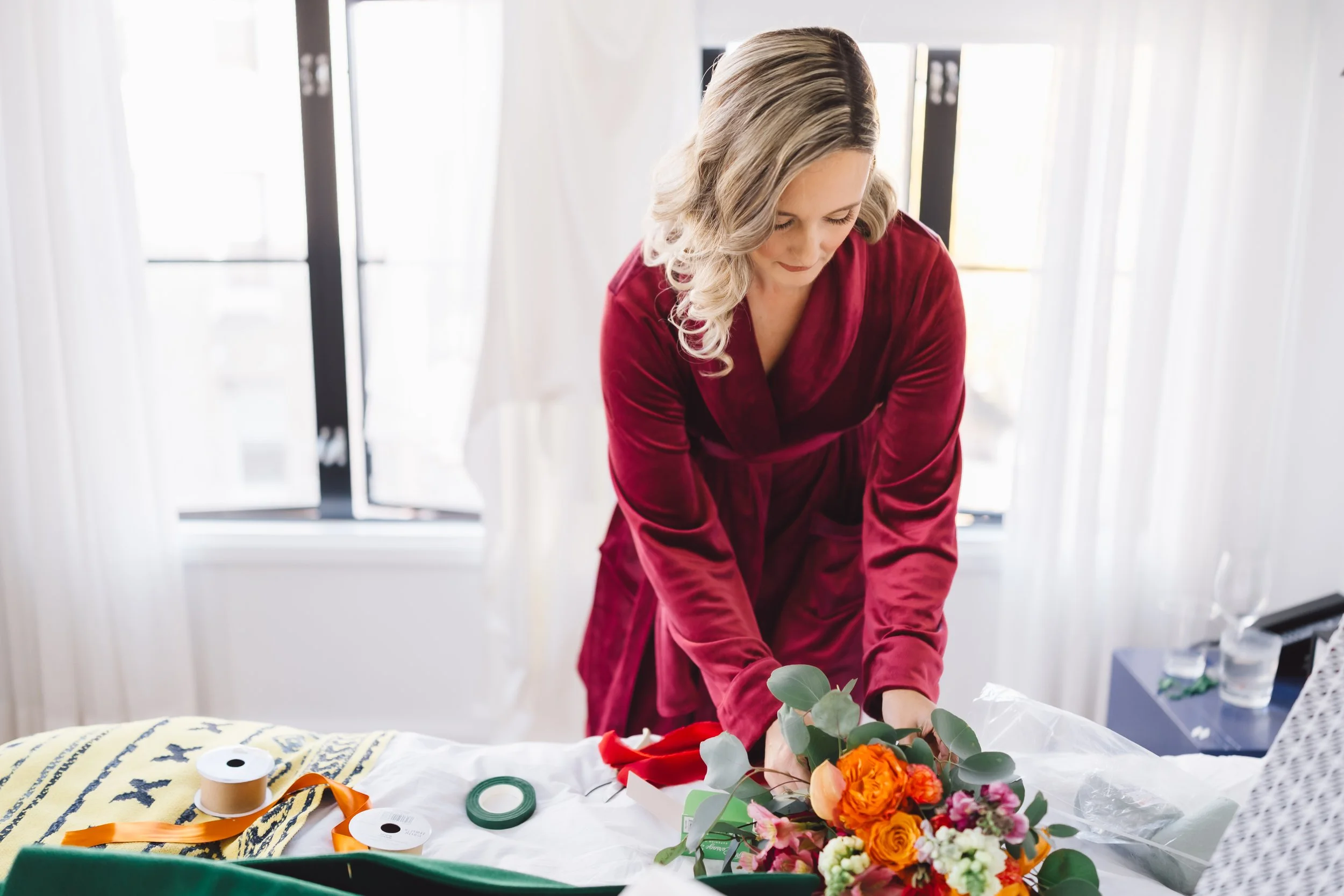 Woman in a red velvet robe arranging flowers on a bed in a bright room.
