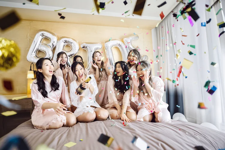 A group of women in pajamas celebrating with confetti and a champagne bottle in a bedroom decorated for a bachelorette party.