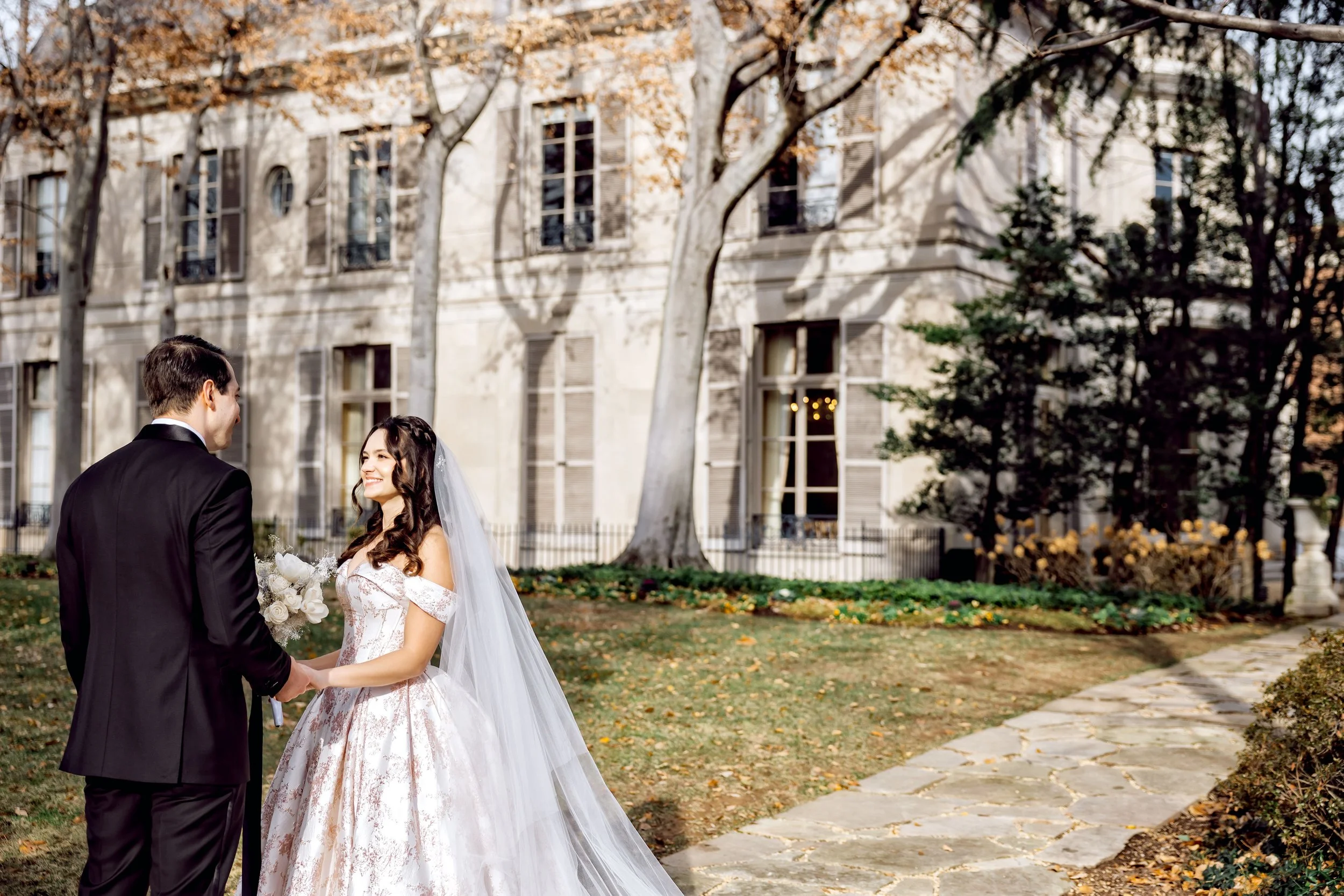 A bride and groom standing outdoors on a park pathway, exchanging vows or rings, with a mansion and trees in the background, during daytime.