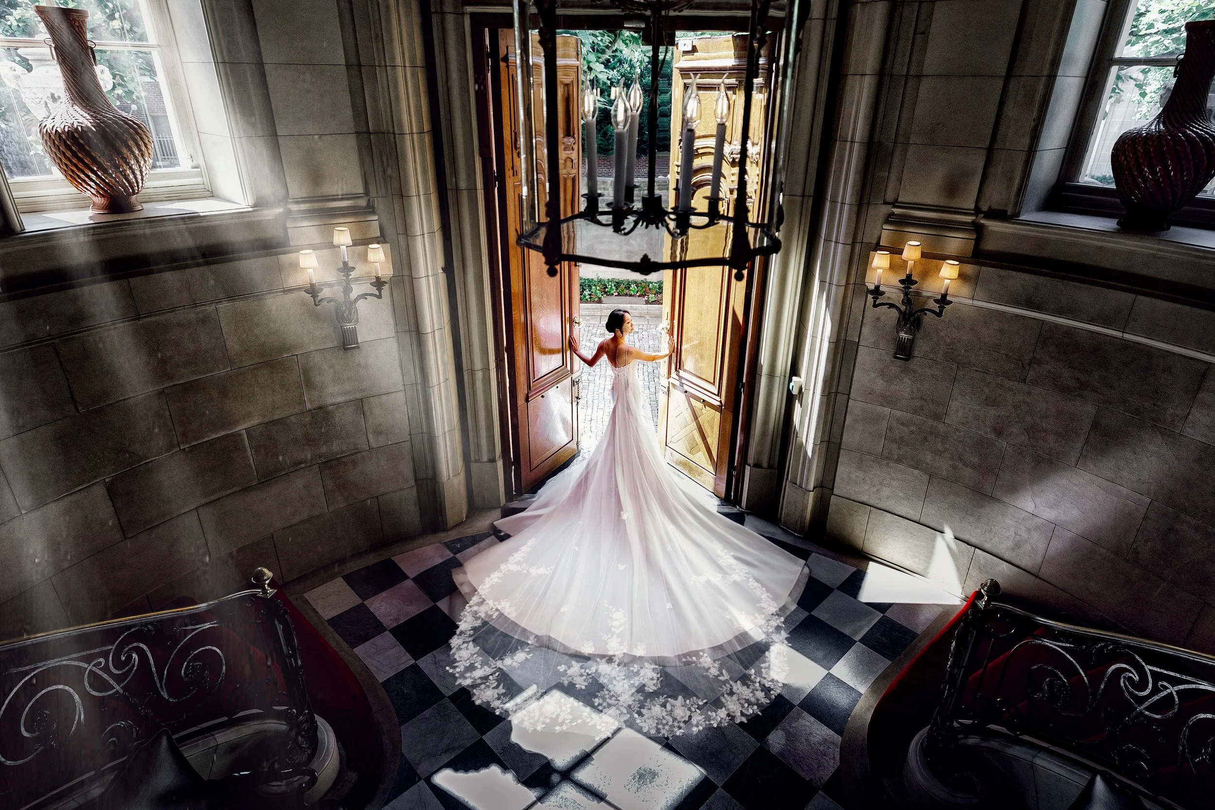 A bride in a white wedding gown stands at the open front door of a grand building, holding the door open with both hands. The gown has a long train and lace details. Bright light is coming from outside, illuminating her and the checkered floor inside