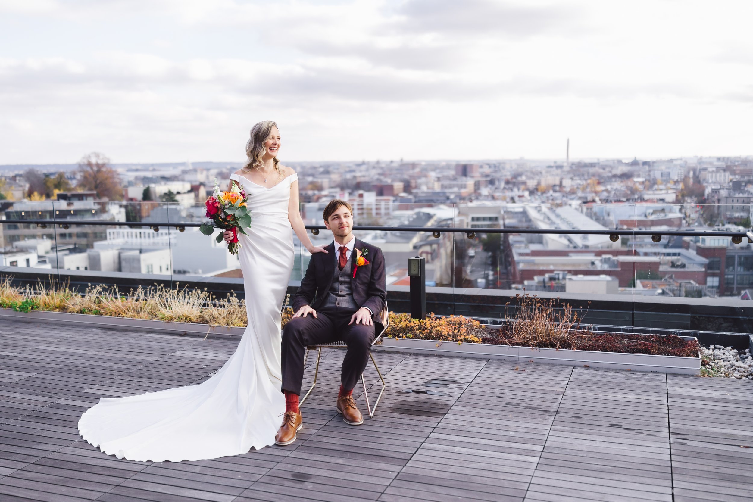 A bride and groom on a rooftop during their wedding photoshoot. The bride is standing and holding a bouquet of colorful flowers, while the groom is sitting on a chair beside her. The bride is wearing a long white wedding dress, and the groom is in a 