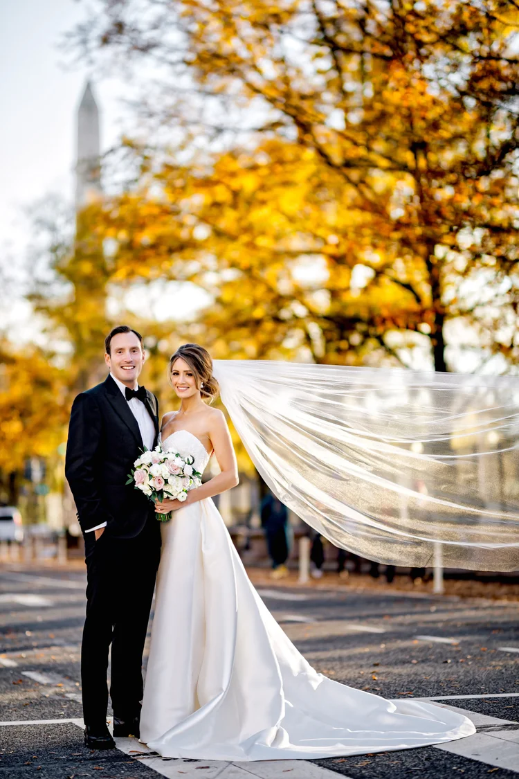 A newlywed couple in wedding attire standing outdoors on a street with autumn trees in the background. The bride is holding a bouquet and her veil is flowing in the wind. There is a tower in the distance.