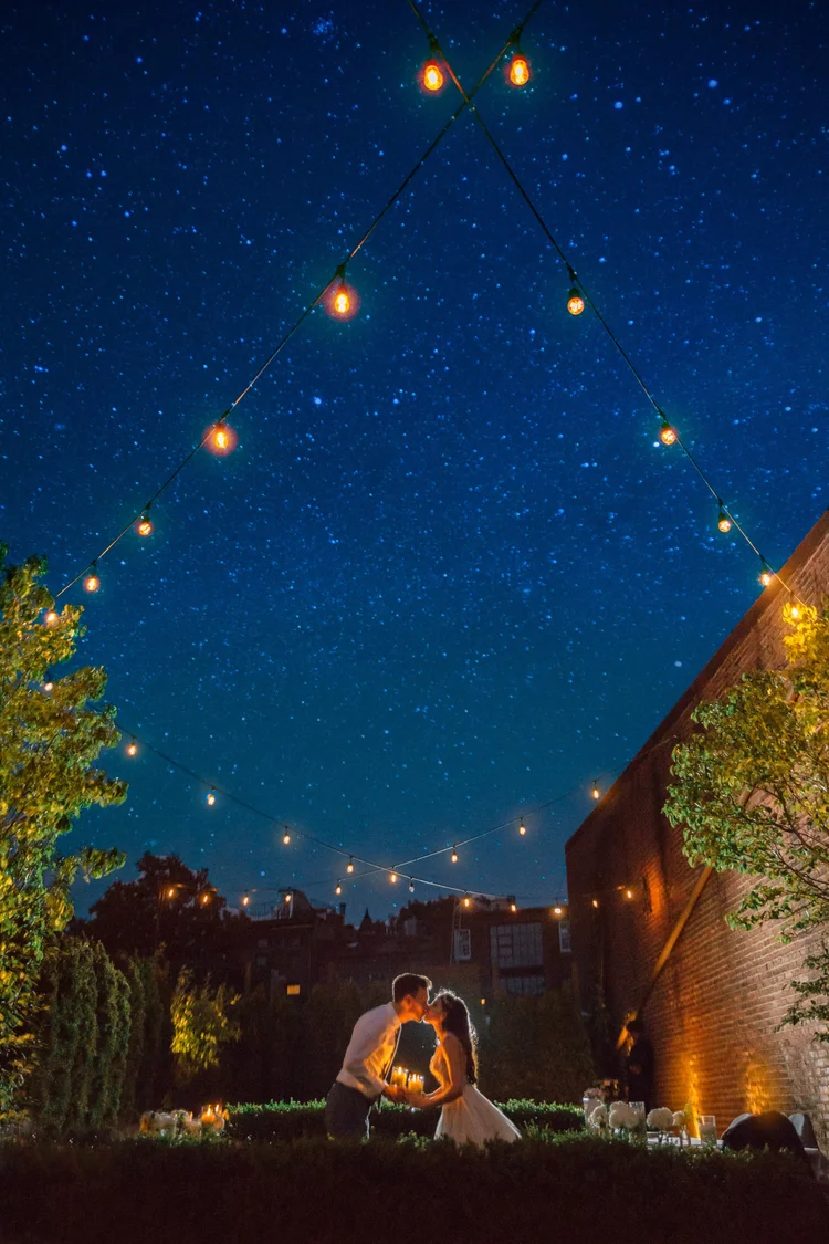 A couple is kissing outdoors at night under string lights, with a starry sky above and greenery around them, illuminated softly for a romantic scene.