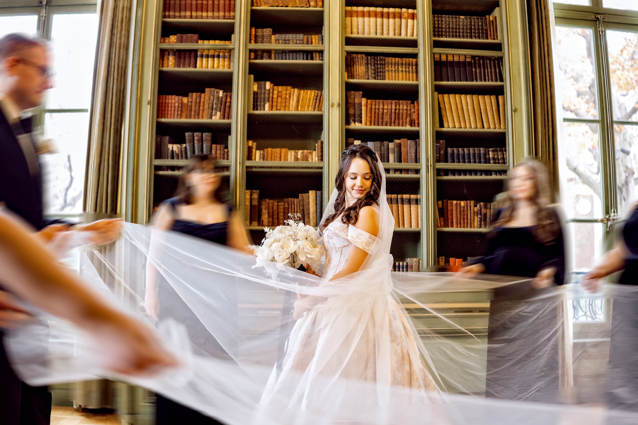 A bride in a white wedding dress holding a bouquet, surrounded by people holding a long veil in a room with large windows and tall bookshelves filled with books.