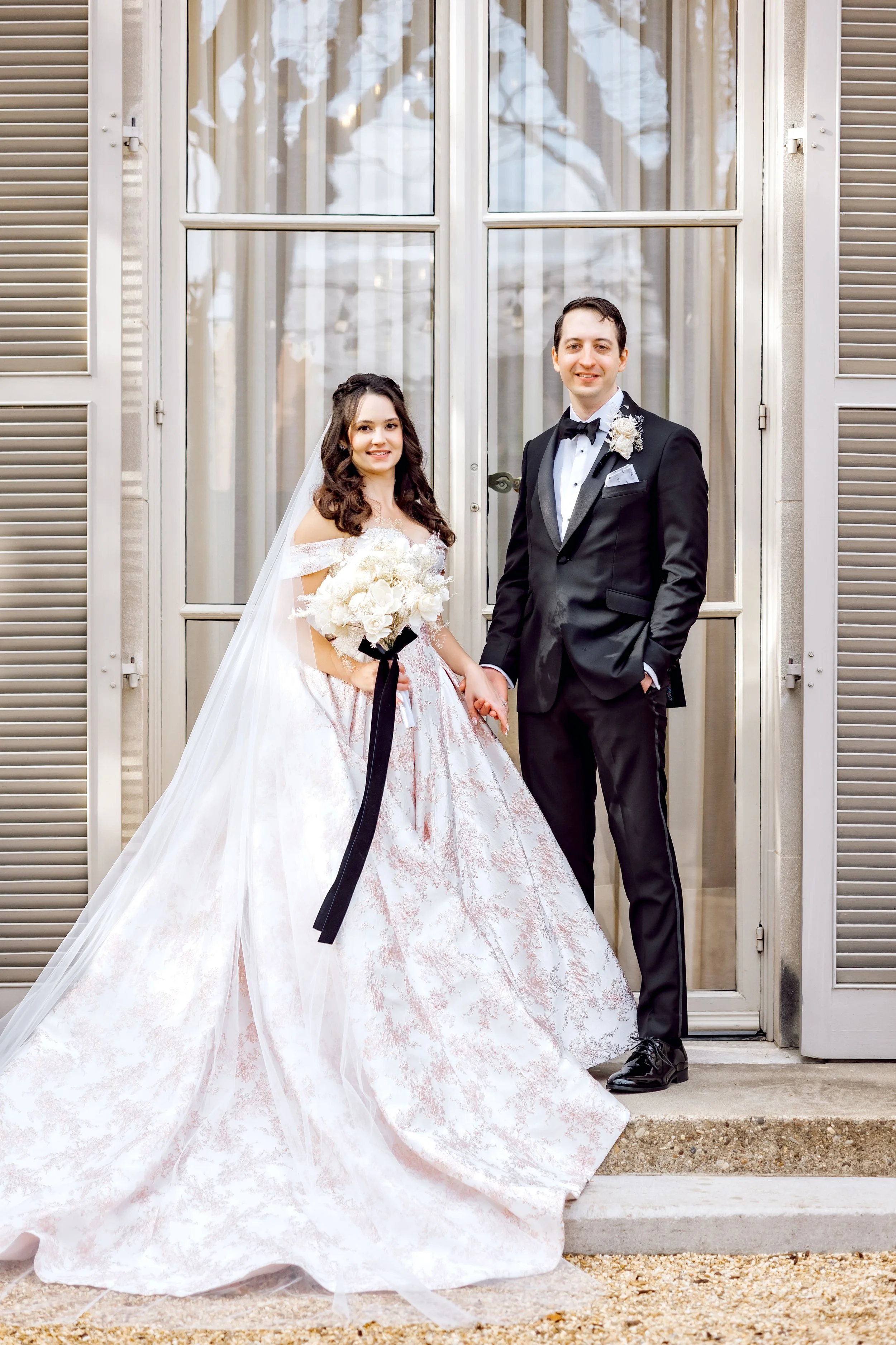 A bride and groom standing hand in hand outside a building with large pane windows and shutters, dressed in wedding attire, smiling at the camera.