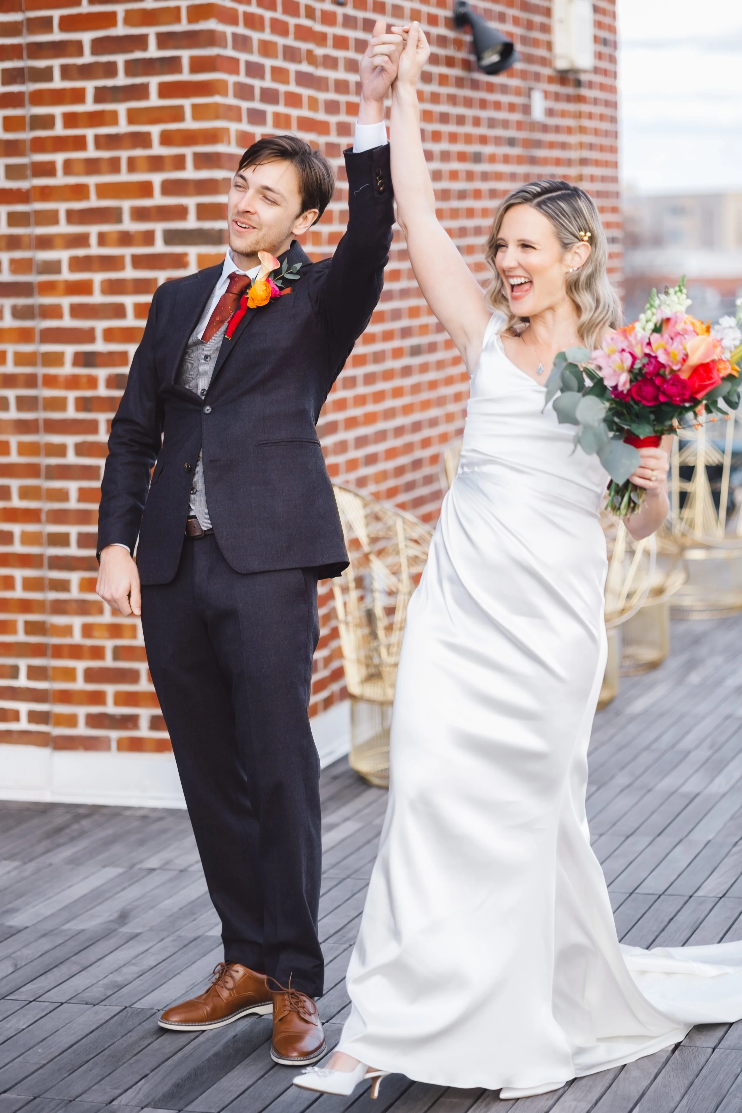 A bride and groom holding hands, celebrating after their wedding ceremony on a rooftop with a brick wall background. The bride is holding a colorful bouquet and is dressed in a white wedding gown. The groom is in a dark suit with a boutonniere.