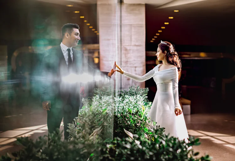 A bride and groom touching hands through a glass wall, in a modern indoor setting with greenery and warm lighting.