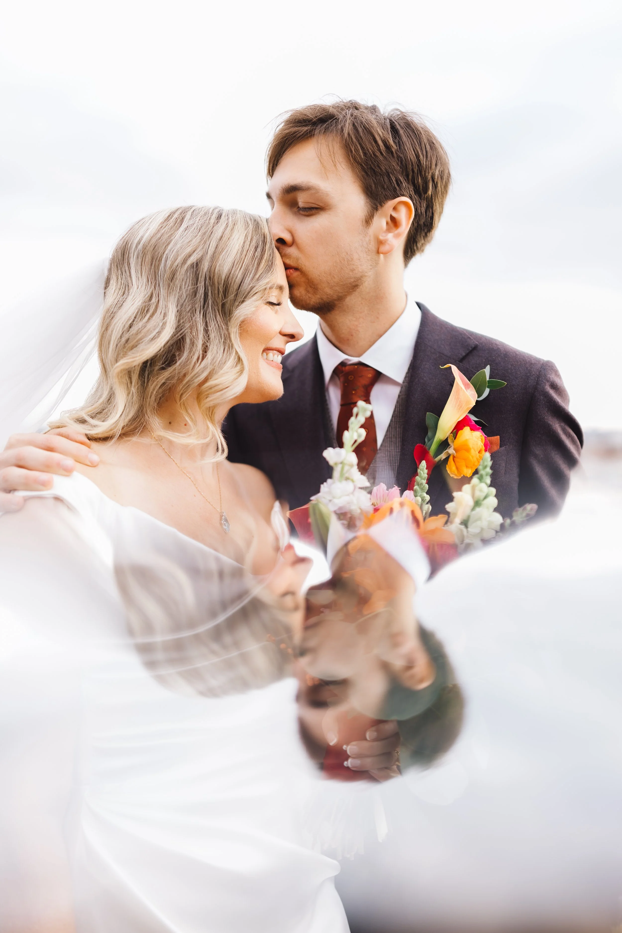 A bride and groom share a tender moment, with the groom kissing the bride on her forehead. The bride has blonde hair and is smiling, holding a bouquet of colorful flowers. The groom is dressed in a suit with a white shirt, brown tie, and boutonniere.