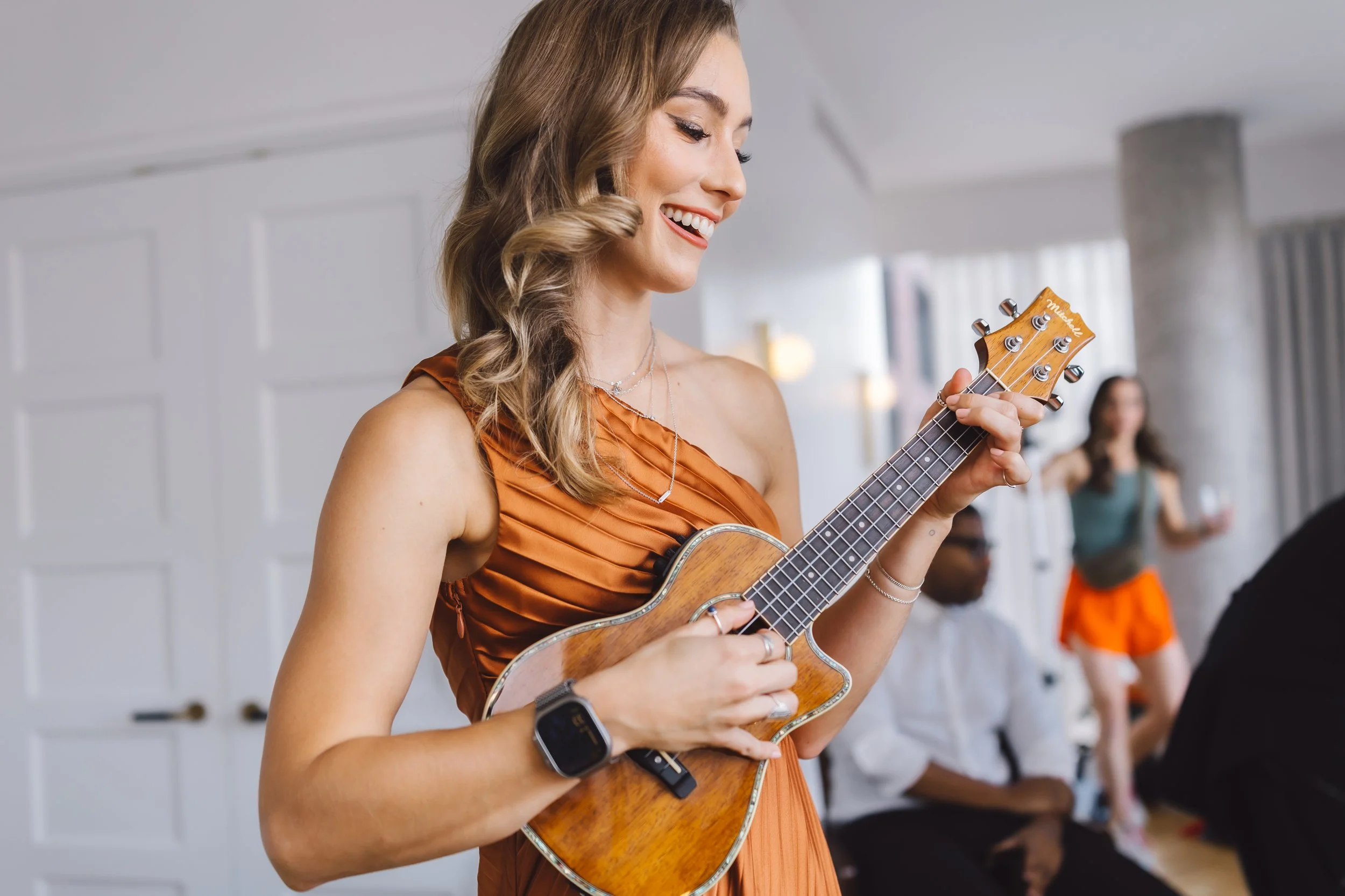 A woman in an orange dress playing a ukulele, smiling during a social gathering indoors. In the background, a woman in a green top and orange shorts holding a drink, and a man in white shirt with suspenders sitting.