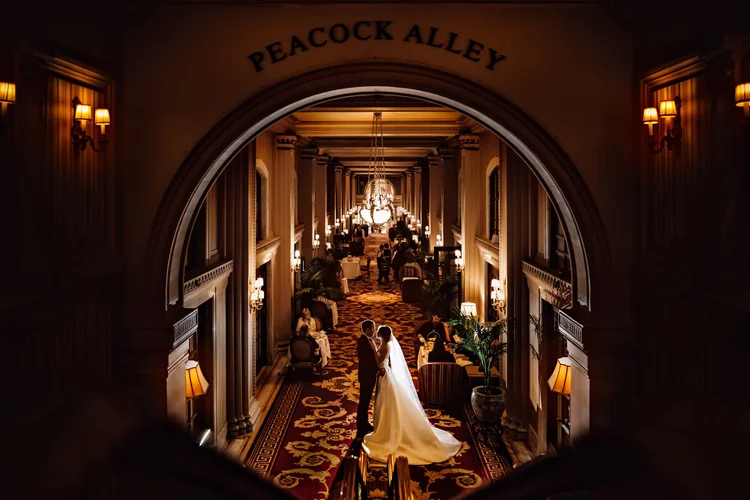 A bride and groom at their wedding in a luxurious, dimly lit hotel hallway with chandeliers and ornate decor, seen through an archway labeled 'Peacock Alley'.