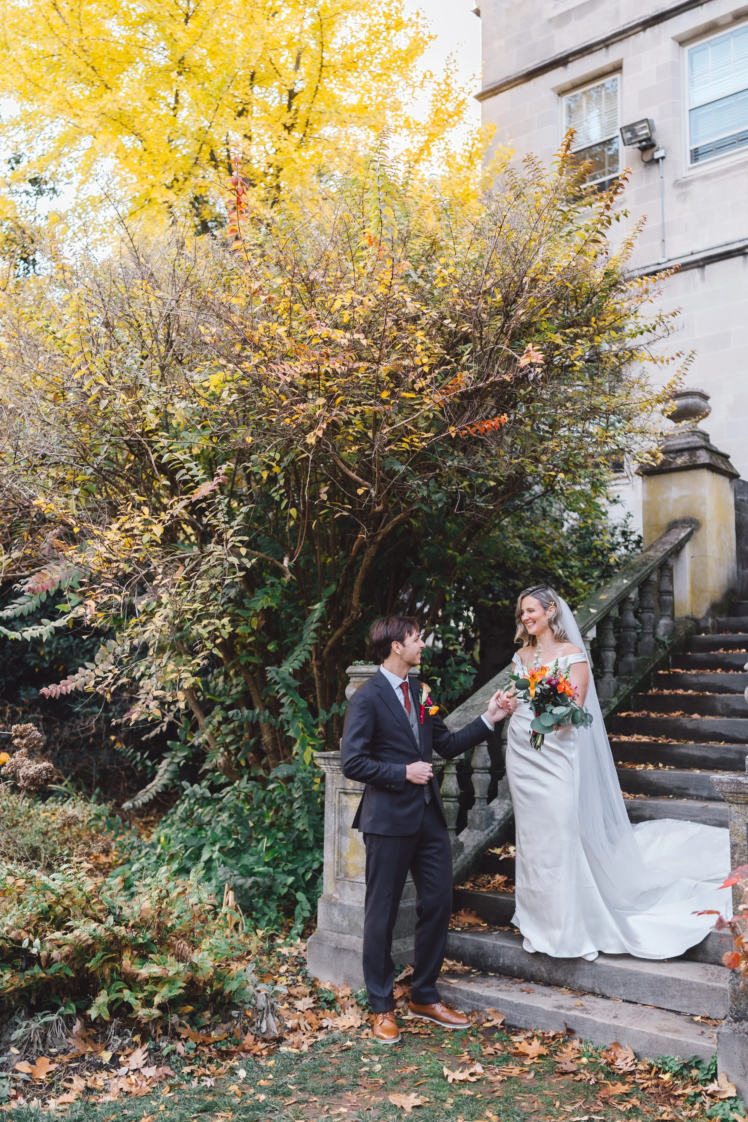 A bride and groom standing on stone steps outdoors during fall, with autumn leaves on trees and ground, and a historic building in the background. The bride holds a bouquet and wears a white wedding dress with a veil, while the groom wears a dark sui