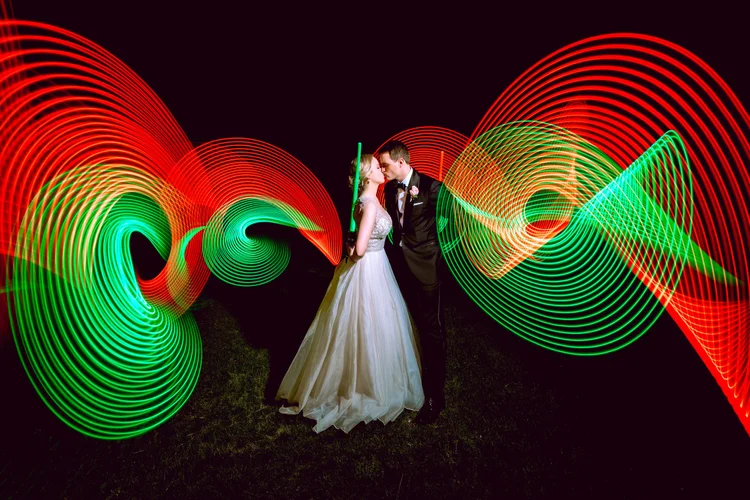 A couple in wedding attire standing close together on a dark background with colorful light trails in red and green swirling around them.
