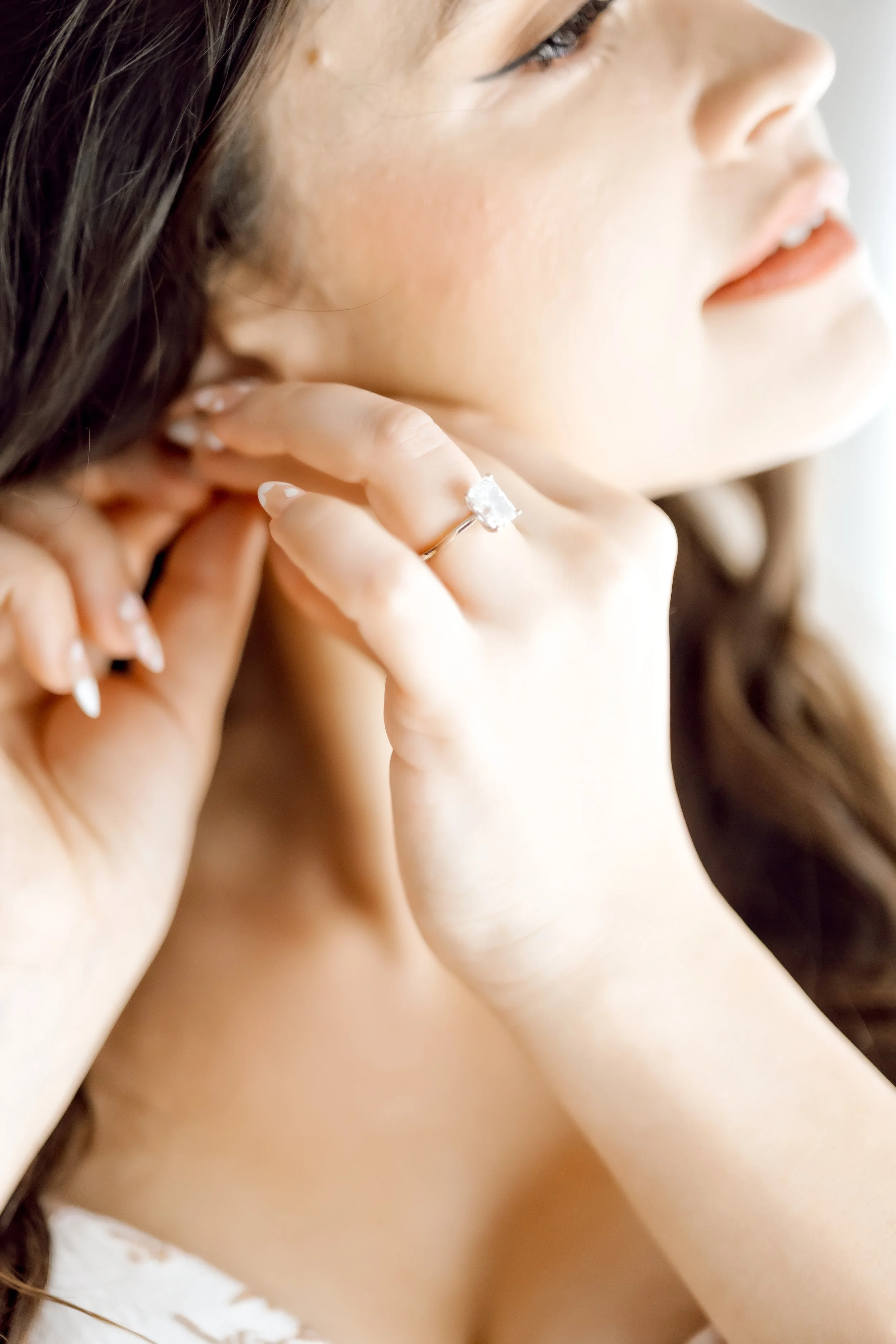 A woman with brown hair wearing a white dress is putting on an earring, with a close-up focus on her face and hand, showing a large diamond ring on her finger.