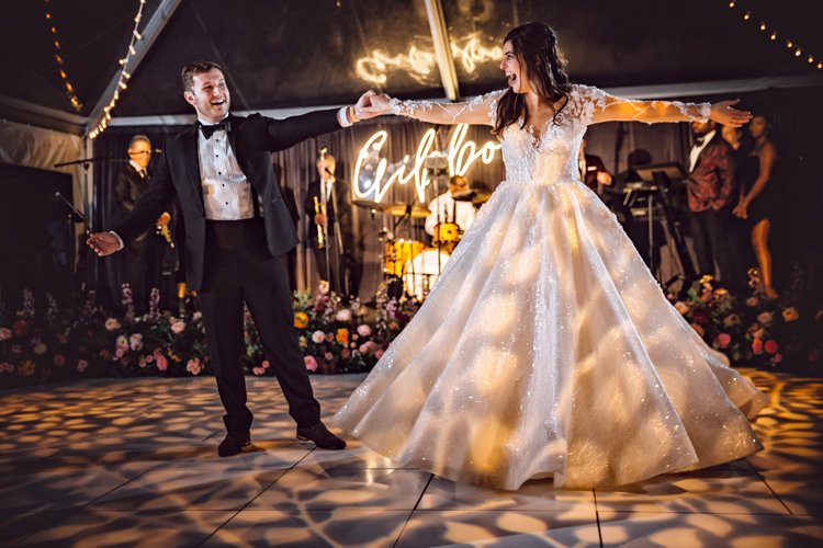 A bride and groom dancing at their wedding reception with a band playing in the background and a neon sign that says 'Guilty'.