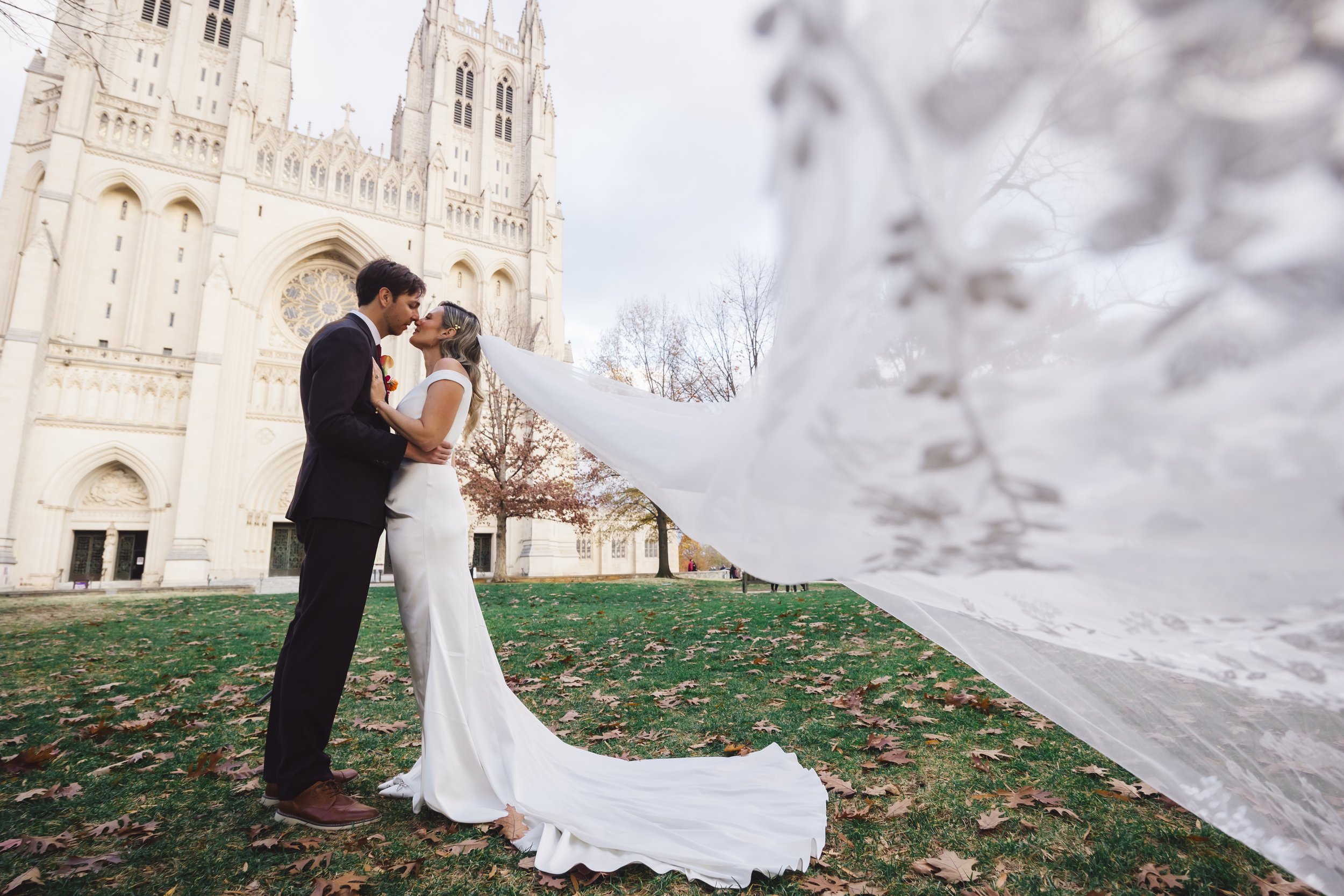 Couple in wedding attire standing close and looking into each other's eyes in front of a cathedral with leafless trees and a fall landscape.