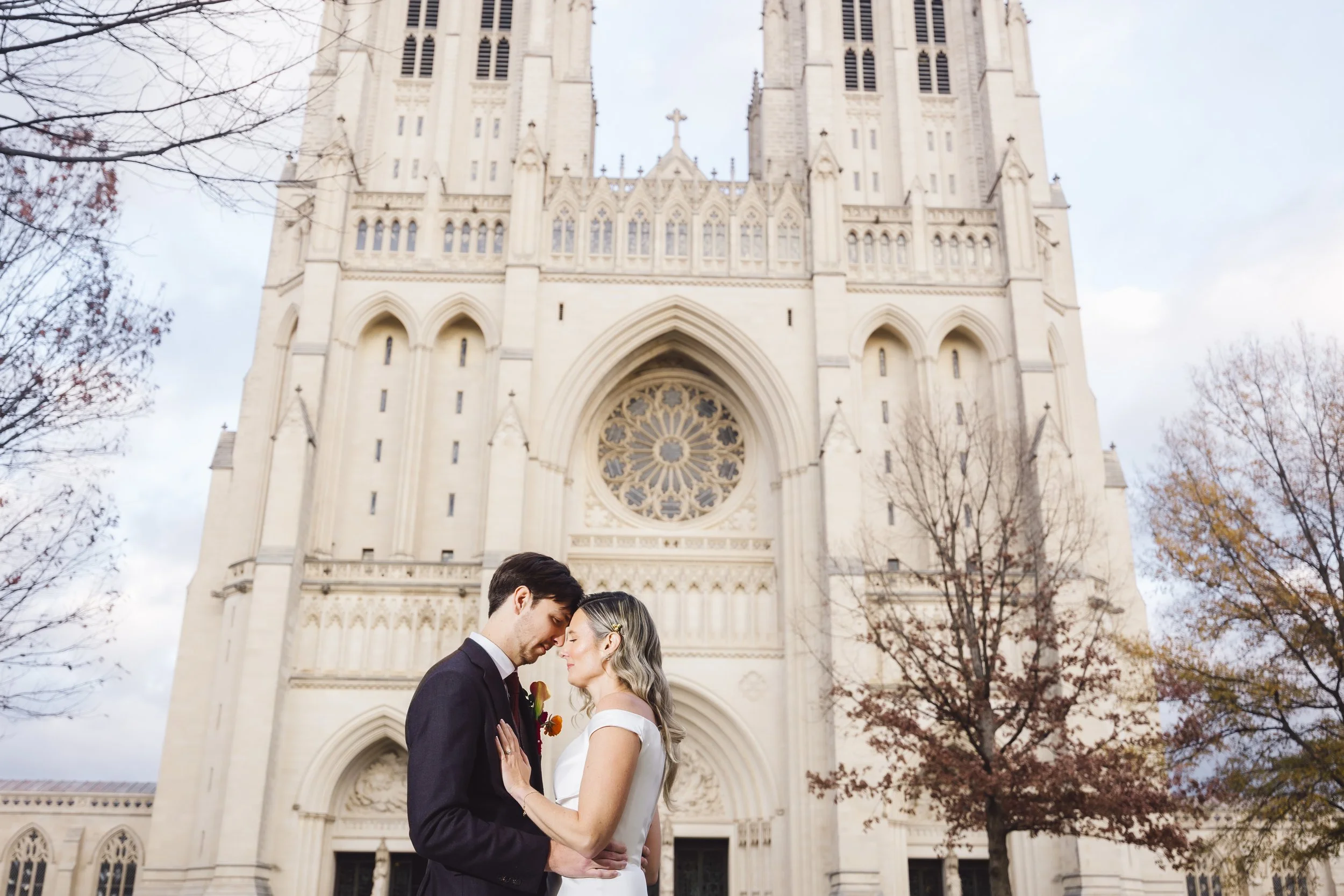 A couple dressed in wedding attire, standing close and touching foreheads in front of a large, historic church with gothic architecture.