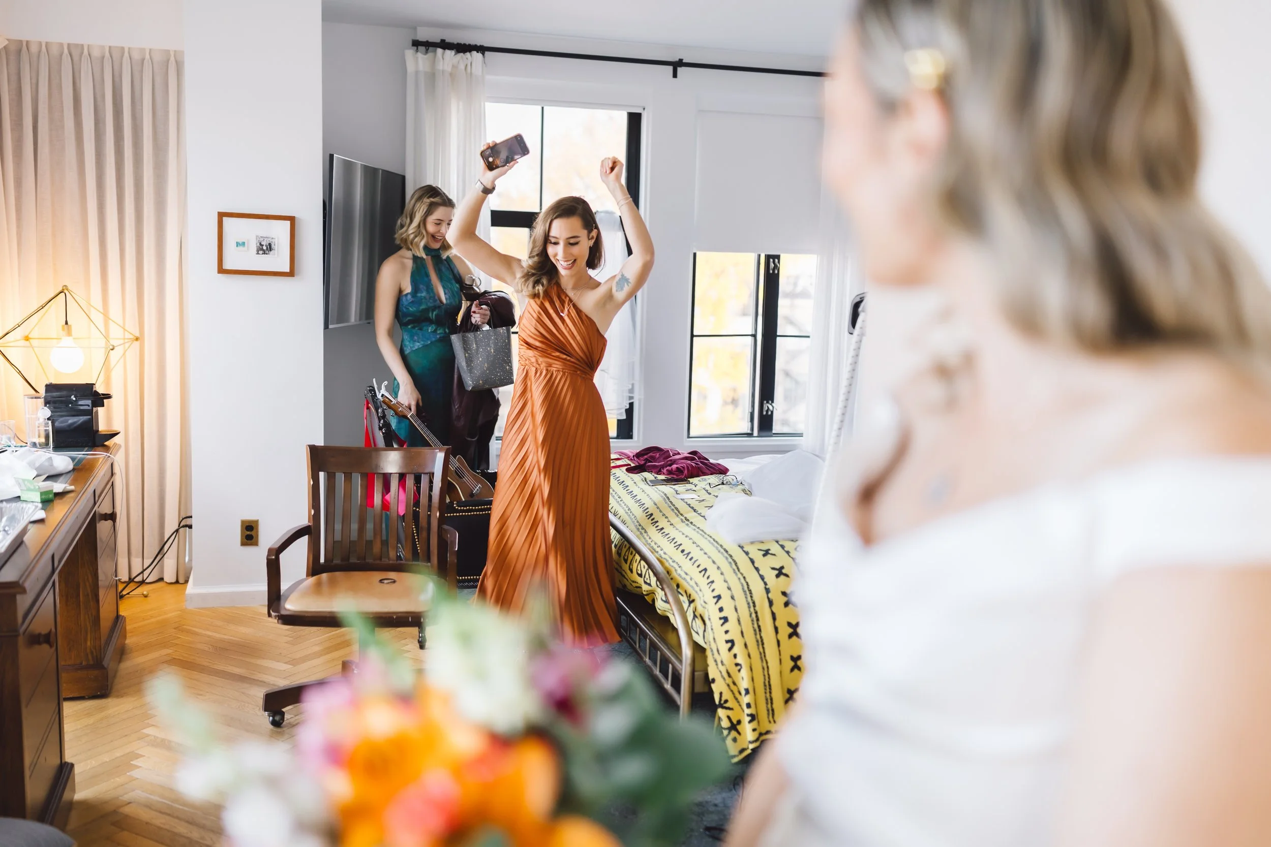 Three women in a brightly lit hotel room, one in a rust-colored dress celebrating, another in a teal dress smiling in the background, and a third woman with gray hair in the foreground, all appearing joyful before a wedding or special event.