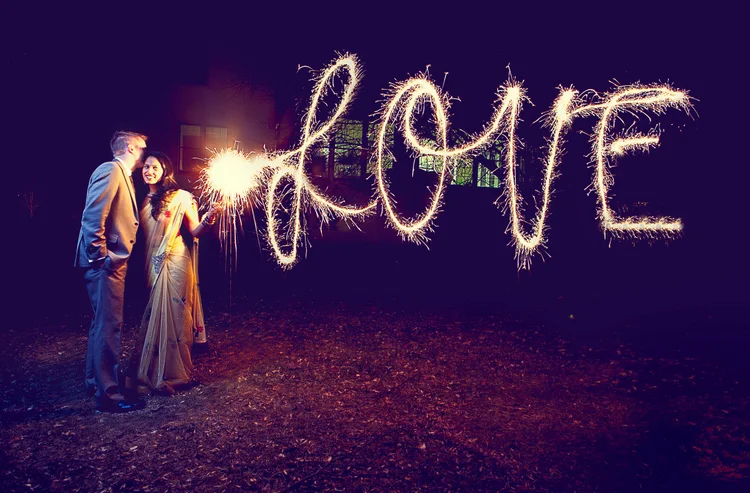 Two people, a man and a woman, standing outdoors at night, holding sparklers with the word 'LOVE' written in sparkler light in the background.