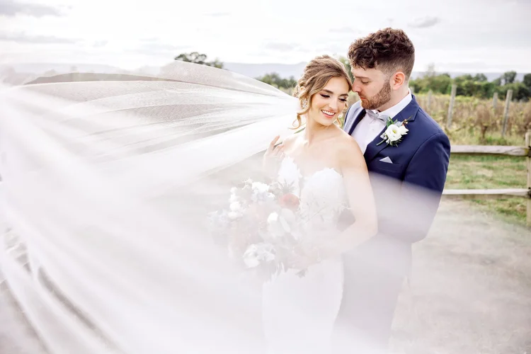 A bride and groom on their wedding day outdoors, smiling and looking at each other, with the bride holding a bouquet and the groom in a navy suit with a boutonniere, and a wedding veil flowing in the wind.