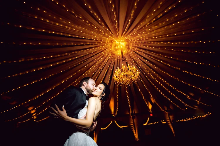A bride and groom sharing a kiss and holding each other in a romantic embrace at their wedding reception under a ceiling decorated with warm string lights and a chandelier.