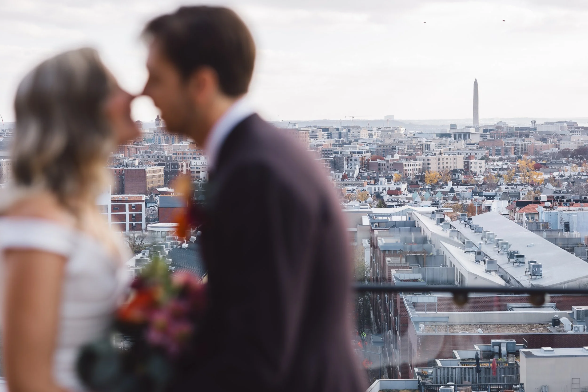 A blurred image of a bride and groom pressing their foreheads together on a rooftop with a cityscape and the Washington Monument in the background.